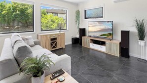 A modern living room with a grey sofa, a coffee table, a TV, and a large window with a view of trees and a house. - The Nook - Lake Illawarra (Lake Illawarra)