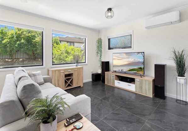 Una sala de estar moderna con un sofá gris, una mesa de centro, un televisor y un ventanal con vistas a árboles y una casa.