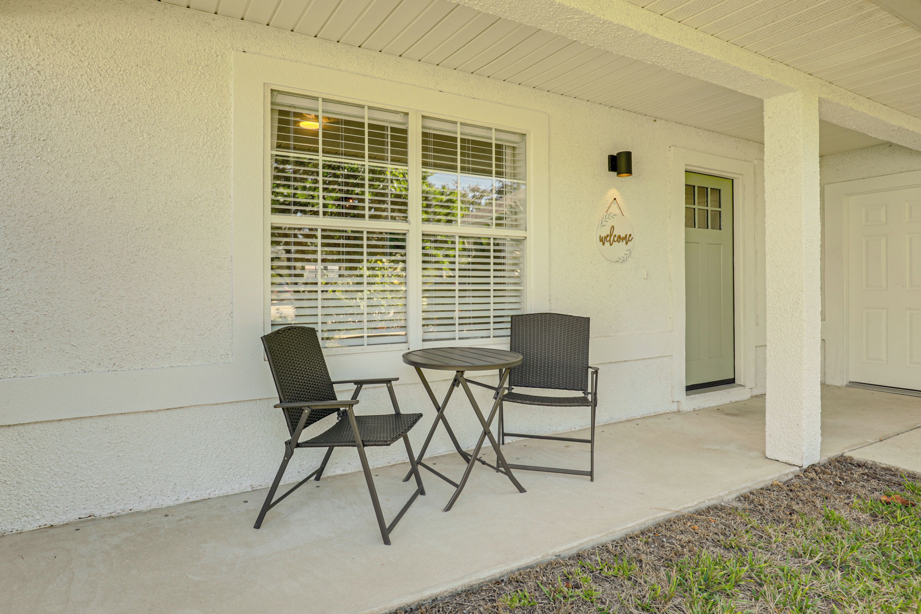 A covered porch with two chairs and a table, a white exterior wall, and a door with a green window.