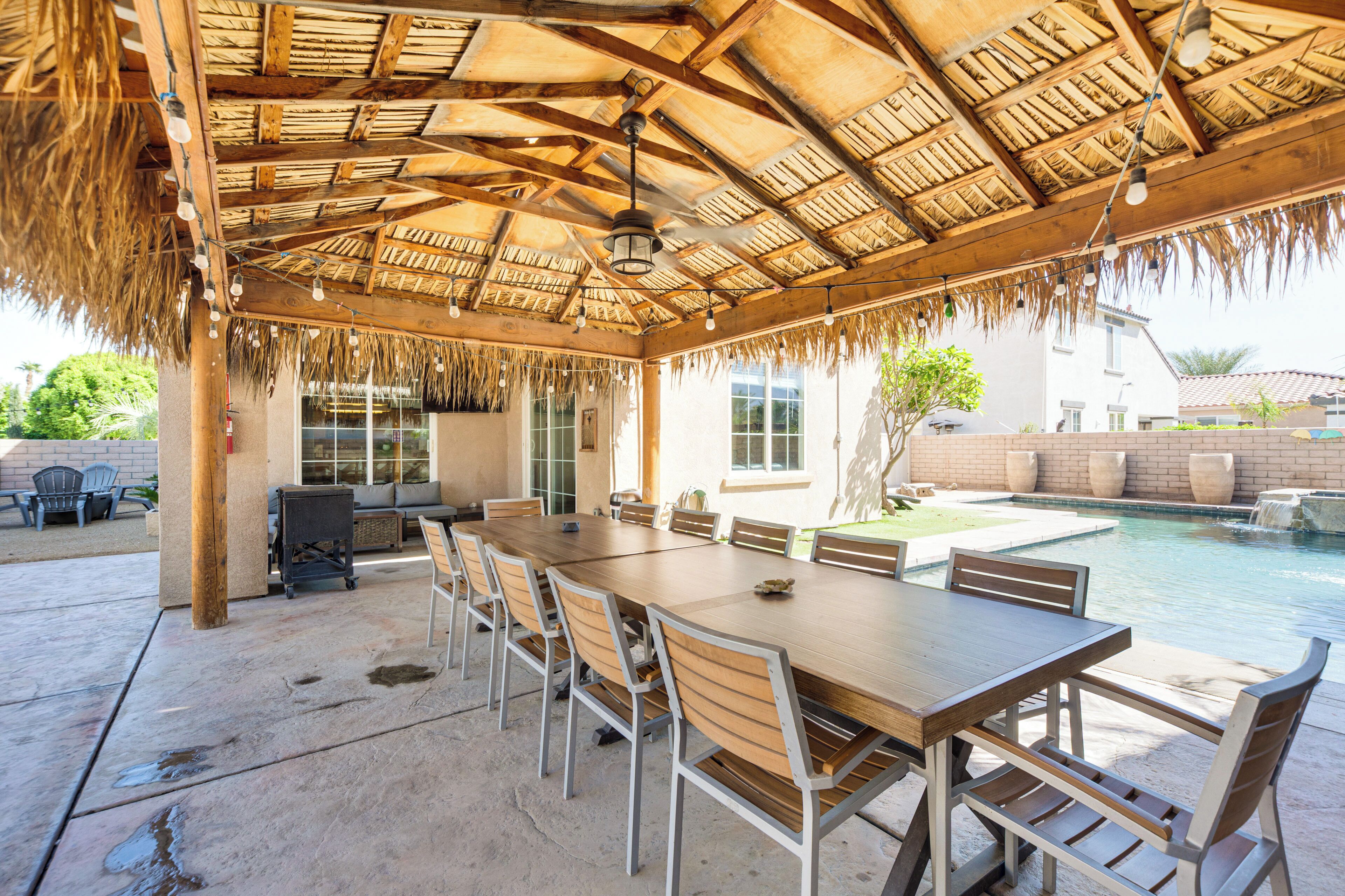 An outdoor covered patio with a dining table and chairs, a pool, and a thatched roof.