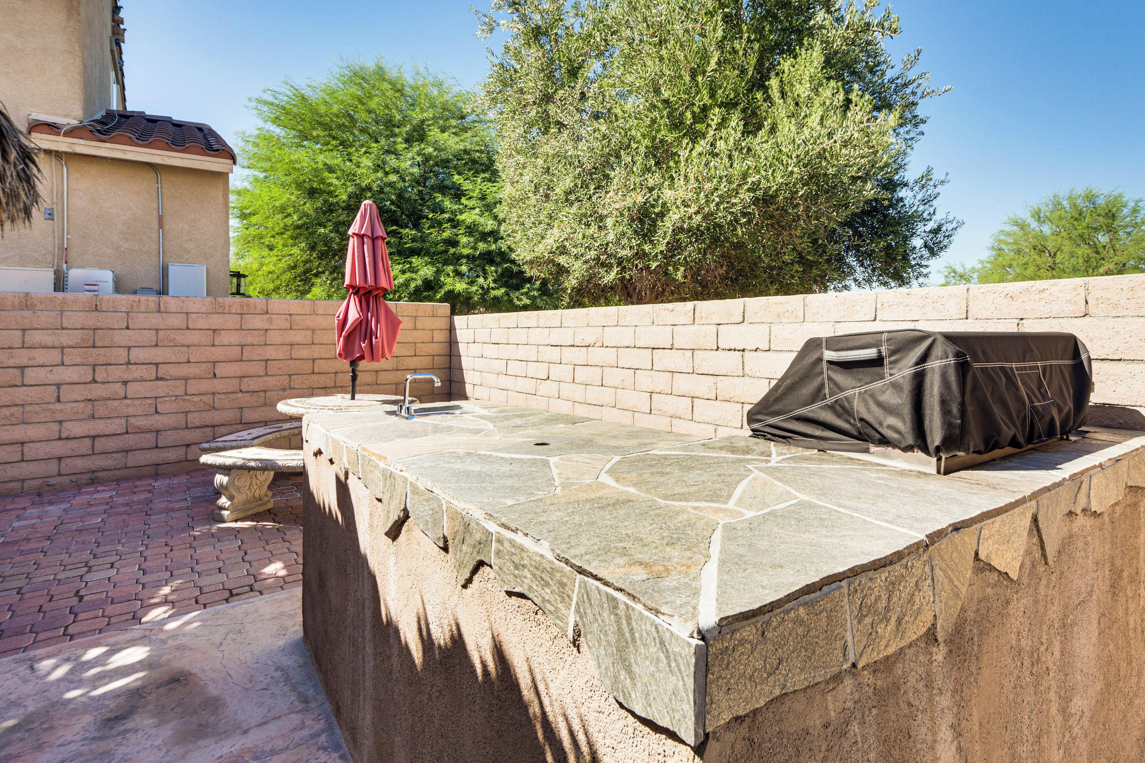 An outdoor patio with a stone table, a grill, and a red umbrella.