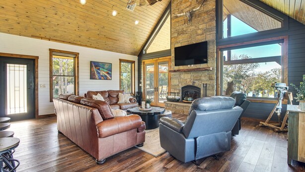 A spacious living room with a fireplace, leather sofas, and a large window offering a view of the outdoors. - Upscale A-frame Escape w/ Lake Access in Quinton! (Eufaula)