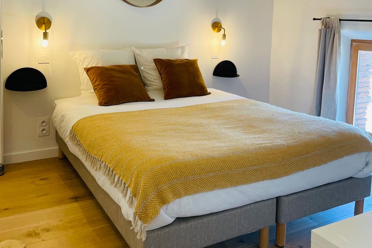 A bedroom with a bed, a round mirror, two wall-mounted lamps, and a window with curtains.