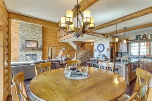A log cabin interior with a dining area featuring a wooden table and chairs, a stone fireplace, and a wooden staircase leading to a loft. - Furnished Decks & Game Room: Sturgis Rally Cabin (Sturgis)