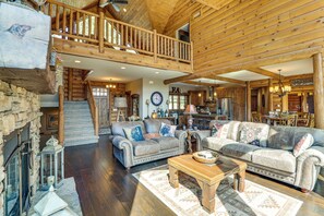 A spacious log cabin interior with a high ceiling, a fireplace, two sofas, a wooden coffee table, and a staircase leading to an upper level. - Furnished Decks & Game Room: Sturgis Rally Cabin (Sturgis)