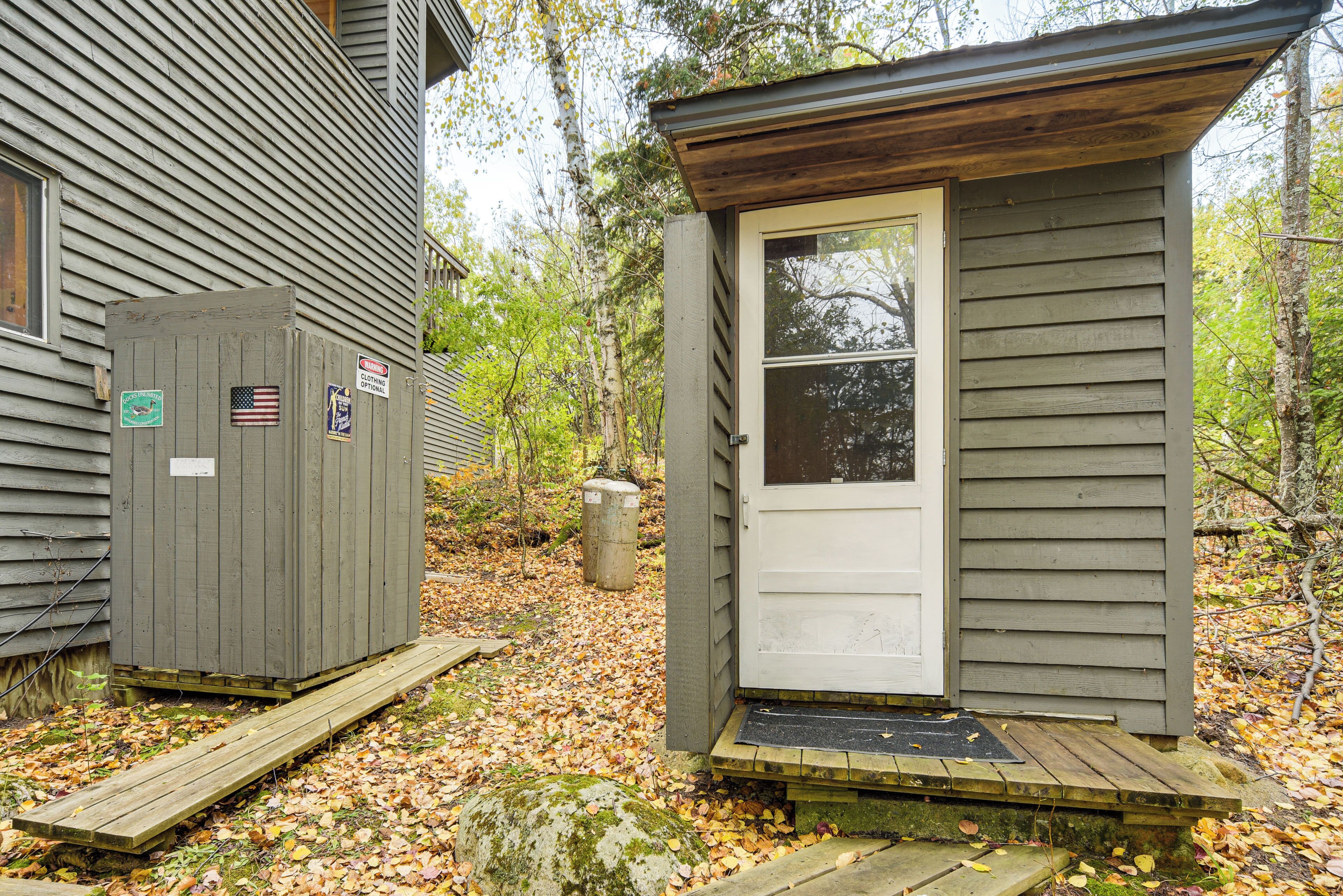 A small wooden cabin with a door and a window, surrounded by trees and fallen leaves.