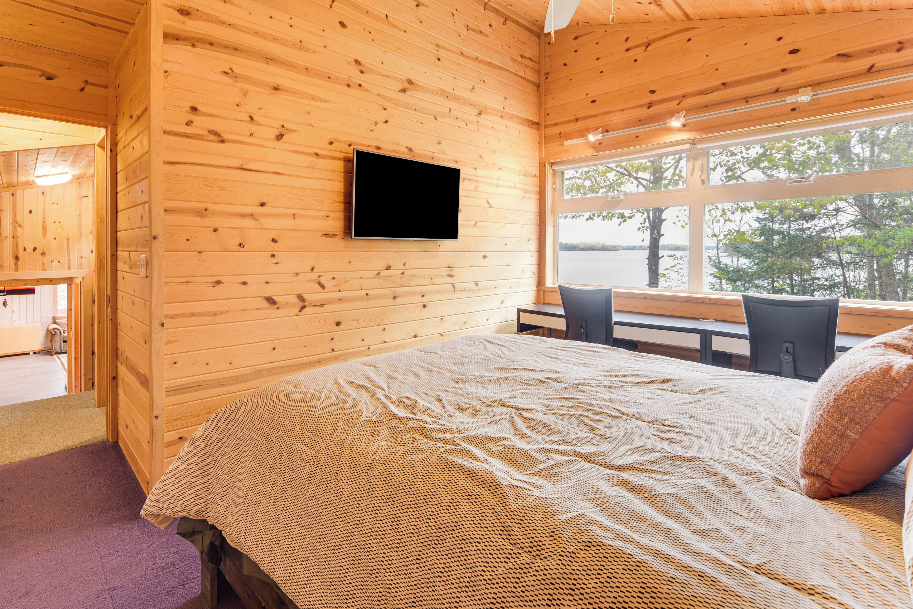 A wooden cabin room with a bed, a TV mounted on the wall, and a window overlooking a body of water.