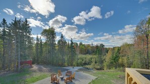 A forested area with a gravel patio, wooden chairs, and a fire pit.