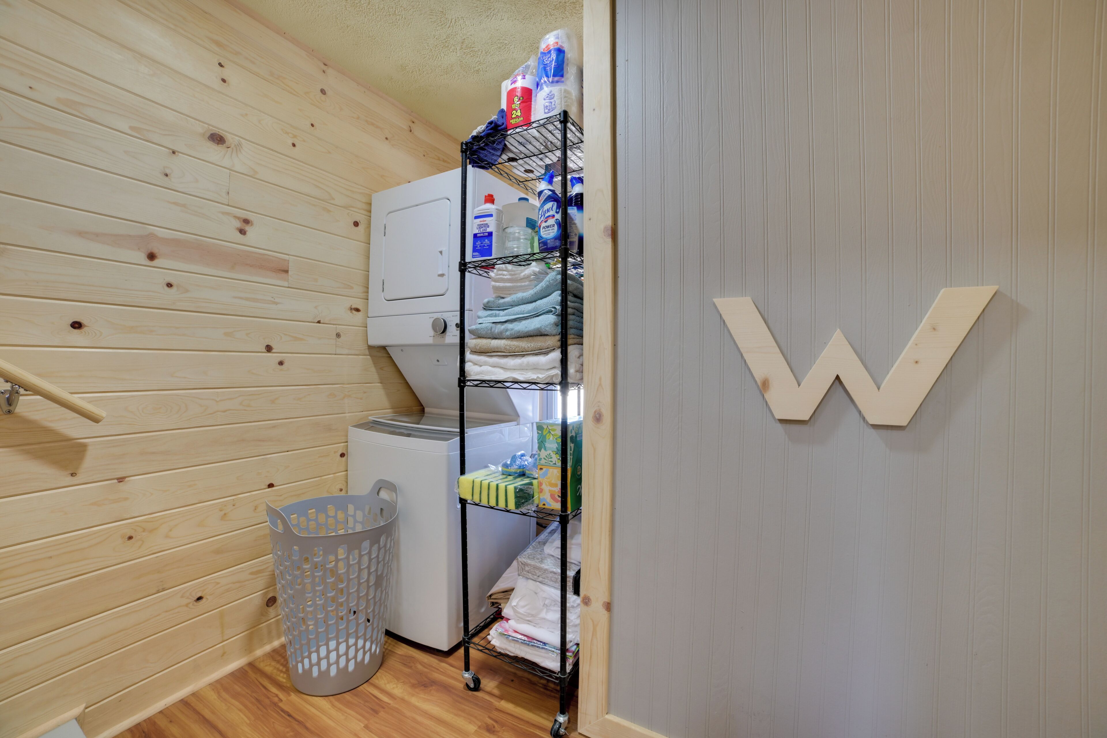 Un lavadero con lavadora, un estante metálico con toallas dobladas, una cesta de ropa y un panel de madera con una letra 'W' grande.