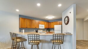 A kitchen with wooden cabinets, a white refrigerator, a microwave, and a clock on the wall. There are three bar stools at the island. - Waterfront Balcony! Condo on Lake of the Ozarks (Lake Ozark)