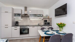 A modern kitchen with white cabinetry, a built-in oven, a microwave, and a coffee maker. There is a dining area with a table set for two, chairs, and a vase of flowers.