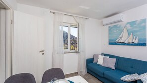 A room with a blue sofa, a dining table with chairs, a window with a view of hills, and an air conditioning unit.