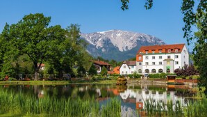 A lakeside view with a mountain in the background, a white building with a red roof, and lush greenery.
