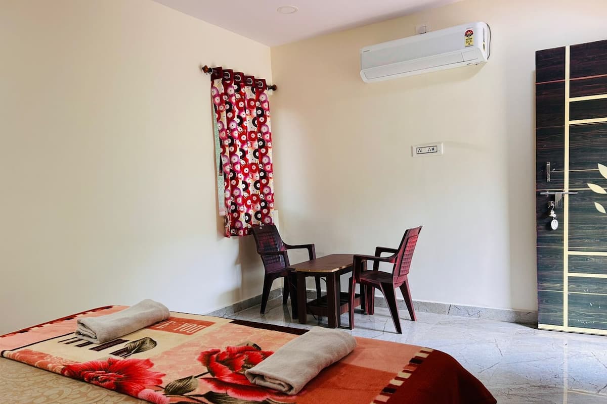 A hotel room with a bed, a small table with two chairs, a window with curtains, an air conditioning unit, and a door with a decorative panel.