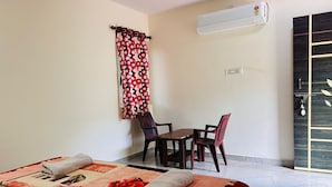 A hotel room with a bed, a small table with two chairs, a window with curtains, an air conditioning unit, and a door with a decorative panel. - Whispering Rocks Jungle Resort (Gangavathi)