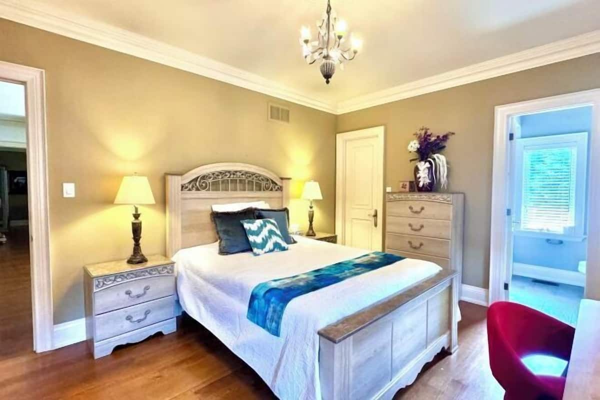 Quarto com cama, mesas de cabeceira, cômoda, lustre e porta que dá para outro espaço.