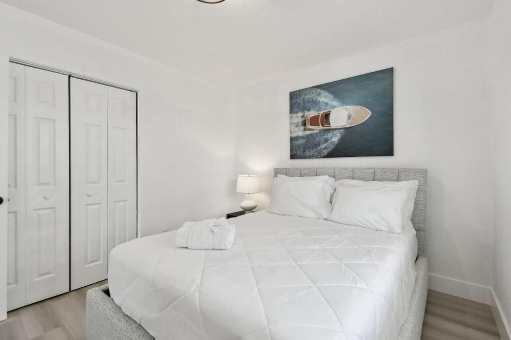 Una cama bien hecha con edredón blanco, una lámpara de noche y un cuadro de un barco en la pared.