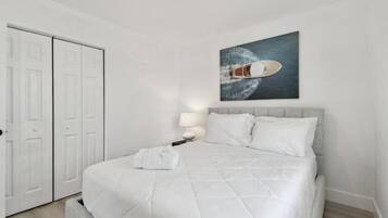 Una cama bien hecha con edredón blanco, una lámpara de noche y un cuadro de un barco en la pared.