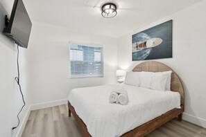 Ein ordentlich eingerichtetes Hotelzimmer mit einem Bett, einem an der Wand befestigten Fernseher, einem Fenster mit Jalousien und einem Bild eines Bootes an der Wand.