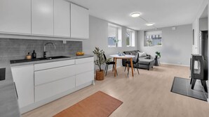 A modern kitchen with white cabinets, a grey backsplash, and a black sink. There is a dining area with a table and chairs, a grey sofa, and a small rug.