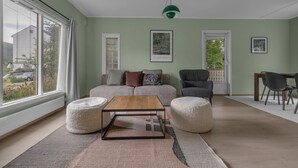 A living room with a sofa, armchairs, a coffee table, and a dining table. There are large windows and a view of the outside.