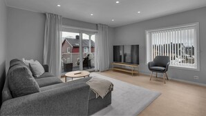 A modern living room with a grey sofa, a TV on a wooden stand, a black chair, and a large window with blinds.