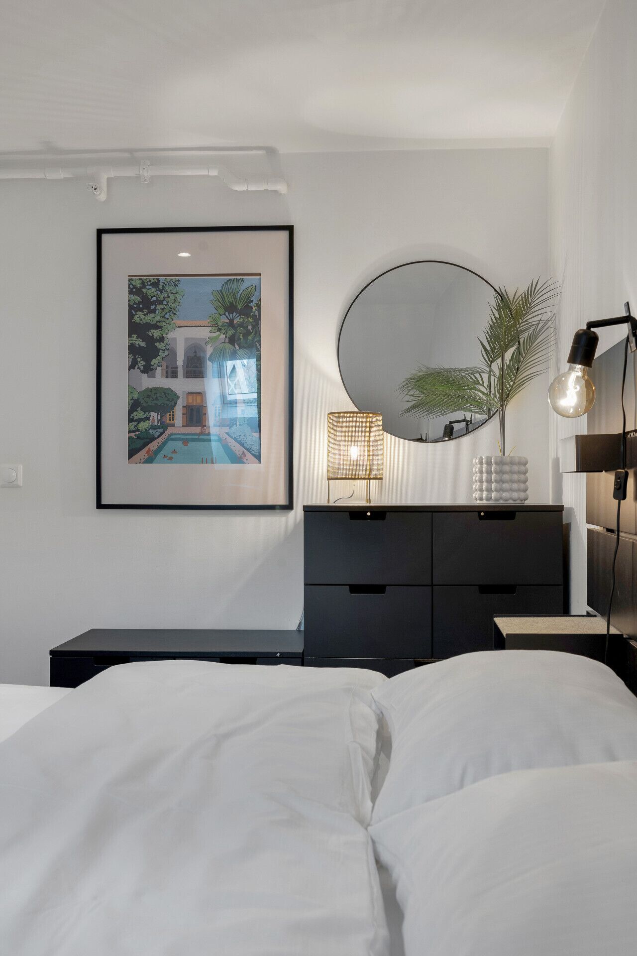 A bedroom with a bed, a dresser, a framed picture, a round mirror, and a lamp.