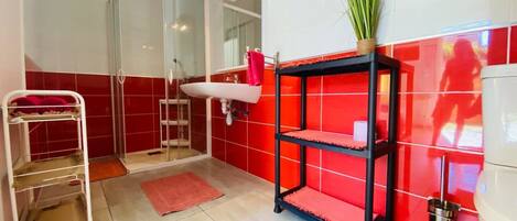 A bathroom with a glass shower enclosure, a white sink, a red tiled wall, a black shelf with a plant, and a white cabinet.