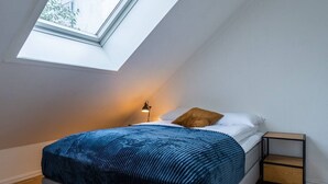 Een slaapkamer met dakraam, een bed met blauwe deken, een bedlamp en nachtkastje.