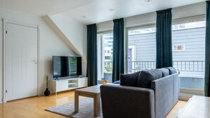 Een moderne woonkamer met een bank, een televisie op een witte kast, een houten tafel en een groot raam met gordijnen.