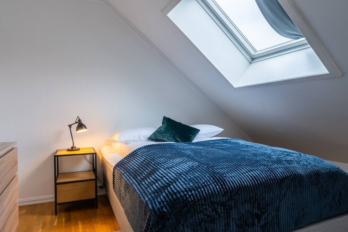 A bedroom with a skylight, a bedside table with a lamp, a bed with a blue blanket, and a wooden dresser.