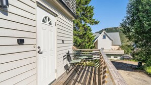 Eine Holzterrasse mit einer weißen Tür, einem kleinen Tisch mit Stühlen und einem Haus im Hintergrund.