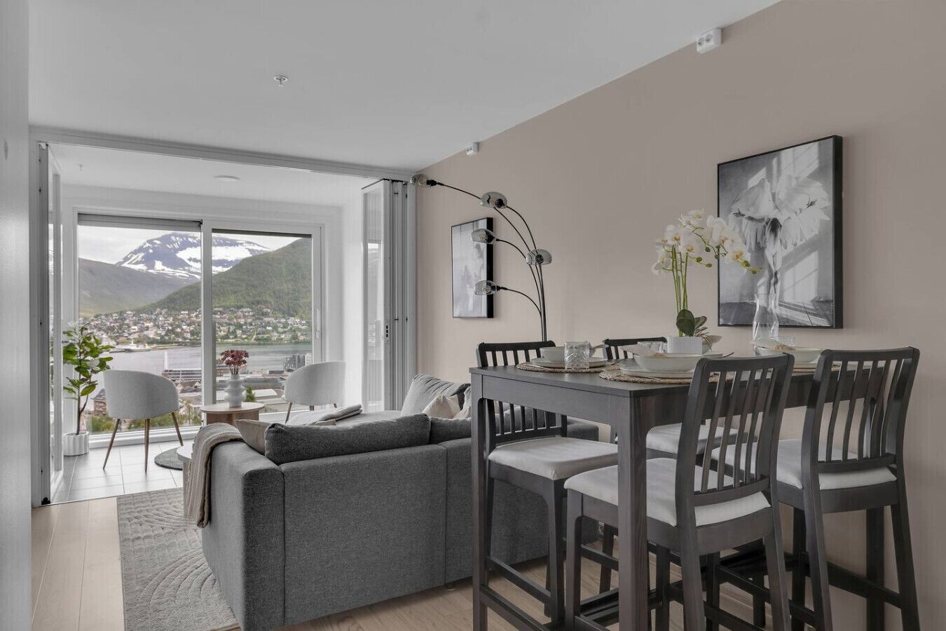 Un salon moderne avec un canapé gris, une table à manger et des chaises. Une grande fenêtre offre une vue sur une montagne et un lac.