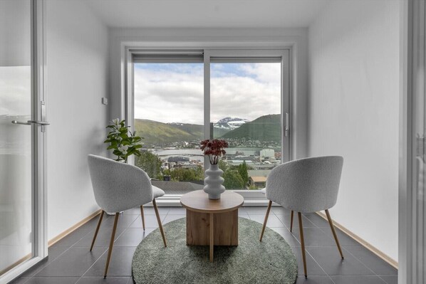 Habitación de esquina con un ventanal grande, dos sillas, una mesa redonda y un jarrón con flores.