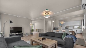 Ein modernes Wohnzimmer mit Flachbildfernseher, grauen Sofas, einem Couchtisch und einem Essbereich mit Küche im Hintergrund.