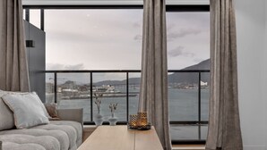 Un salon moderne avec un canapé, une table basse en bois et une vue sur le paysage urbain et l’eau, grâce à de grandes fenêtres.