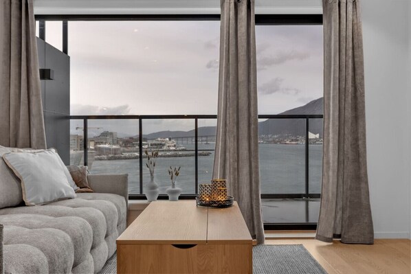 A modern living room with a sofa, a wooden coffee table, and a view of a cityscape and water through large windows.