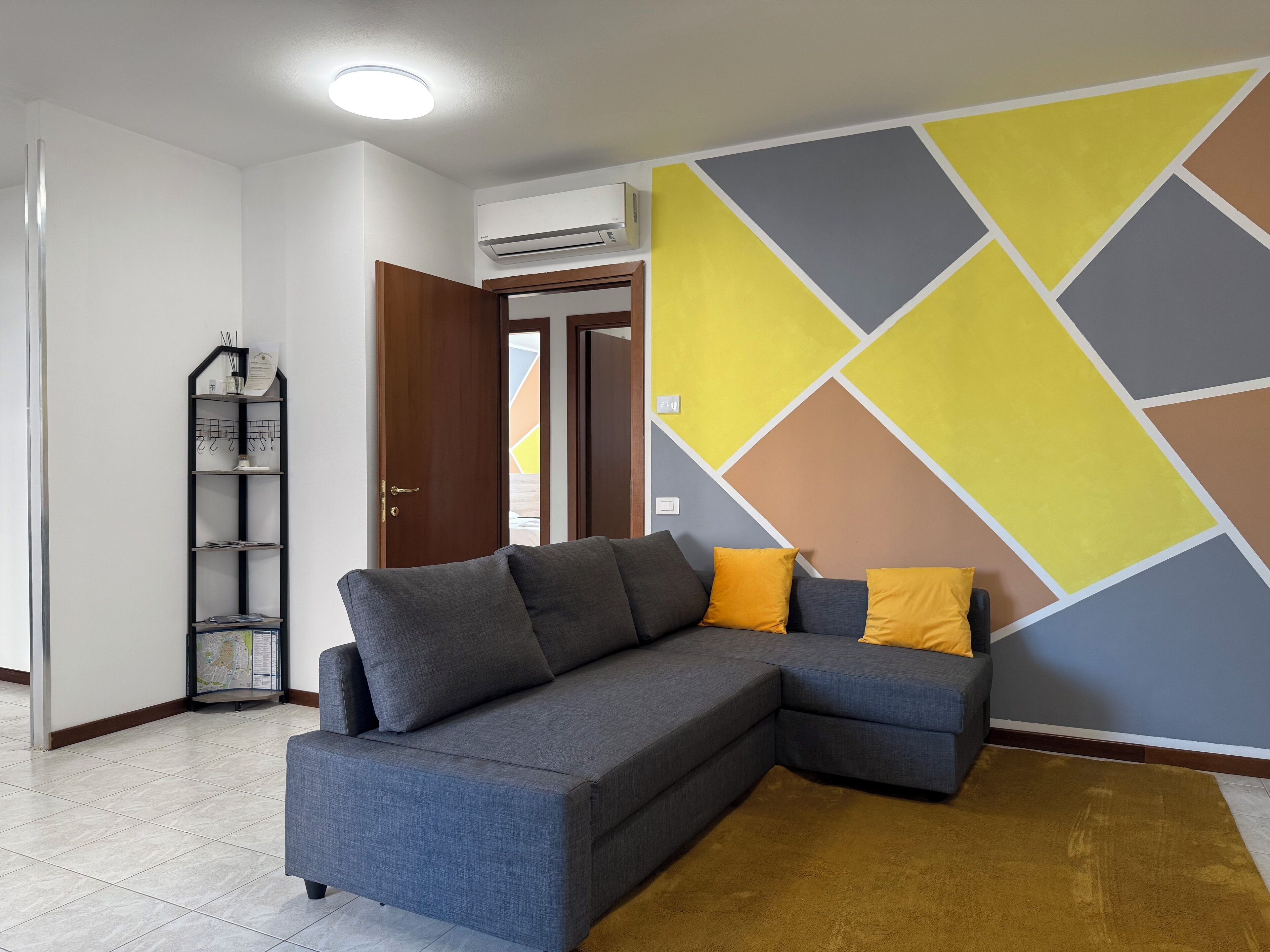 Ein modernes Wohnzimmer mit einem grauen Ecksofa, einem gelb-grauen geometrischen Wanddesign, einem braunen Teppich und einem kleinen schwarzen Regal mit Gegenständen.