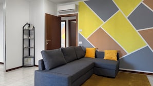 A modern living room with a grey sectional sofa, a yellow and grey geometric wall design, a brown rug, and a small black shelf with items.