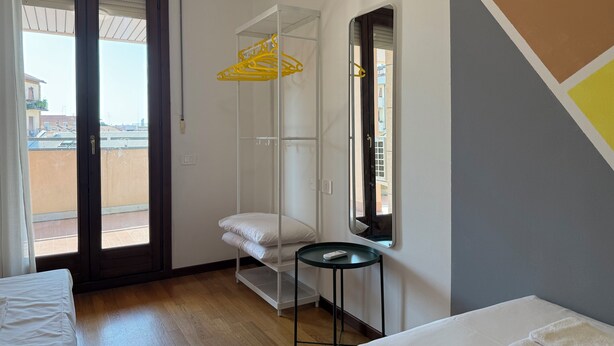 A hotel room with a bed, a small table, a window with blinds, and a balcony view.