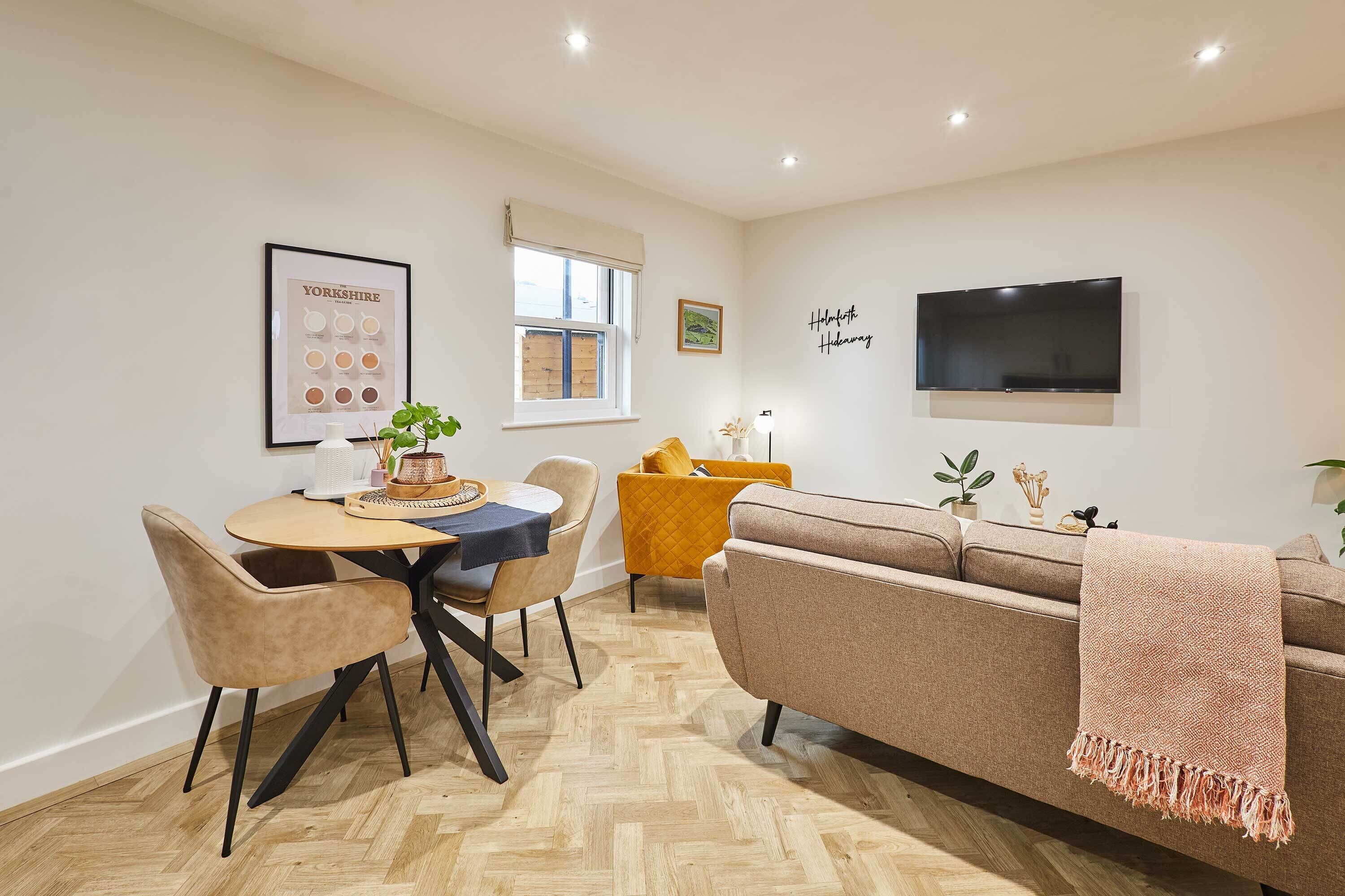 A modern living room with a sofa, a dining table with chairs, a TV mounted on the wall, and a window with a view.