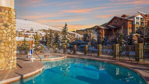 A pool area with a stone wall, a hot tub, and a view of a residential area with snow-covered roofs and trees. - Grand Lodge Crested Butte | King Suite, Walk to Lifts, Pool & Hot Tub, View (Crested Butte)