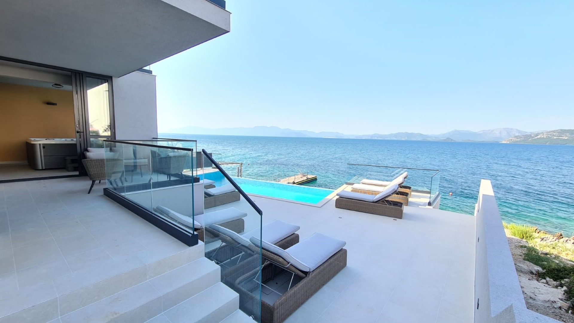 A modern outdoor terrace with a glass balustrade, a pool, and lounge chairs overlooking the sea.