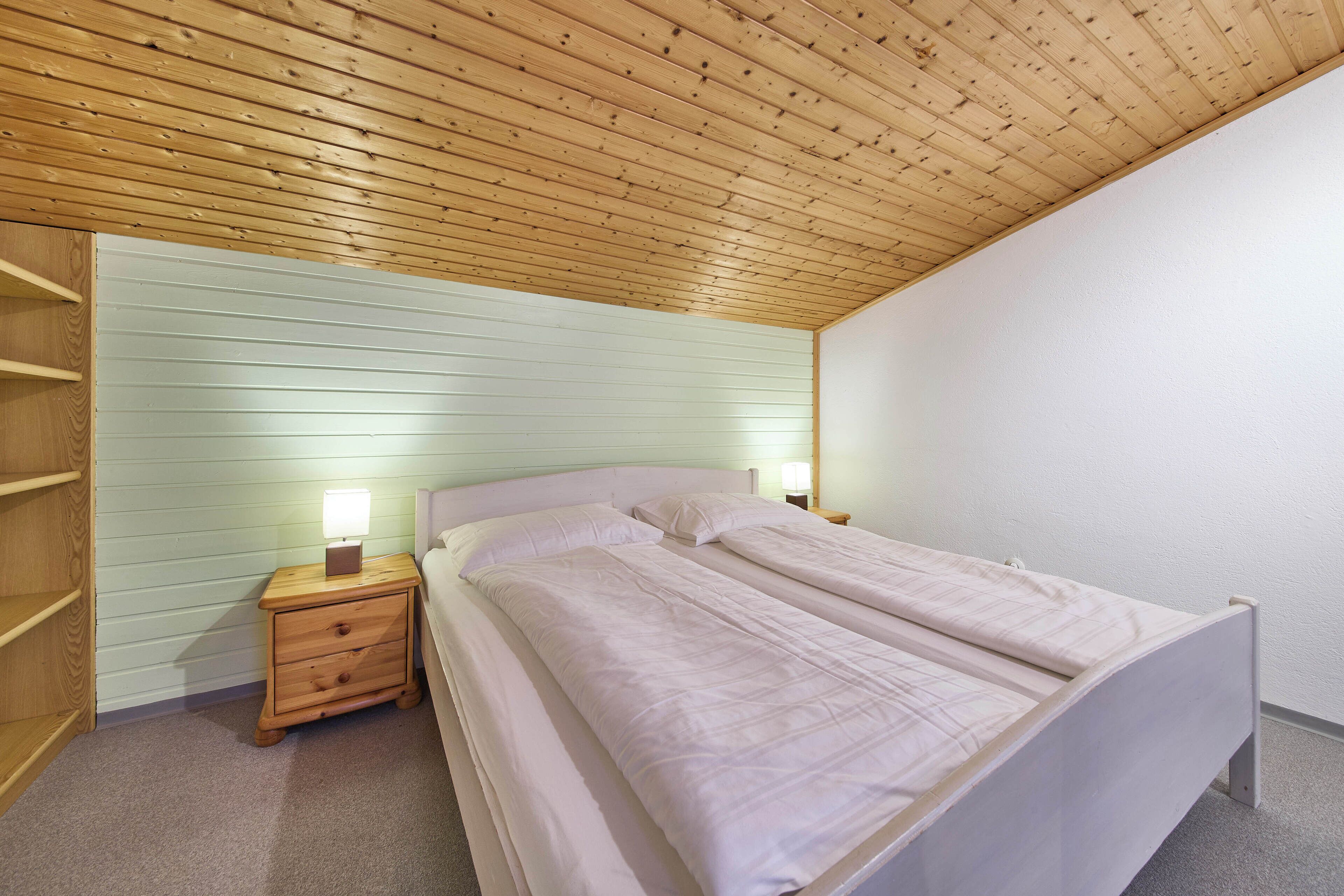 Une chambre avec un plafond en bois, un lit avec une literie blanche, une table de chevet avec une lampe et une étagère en bois.