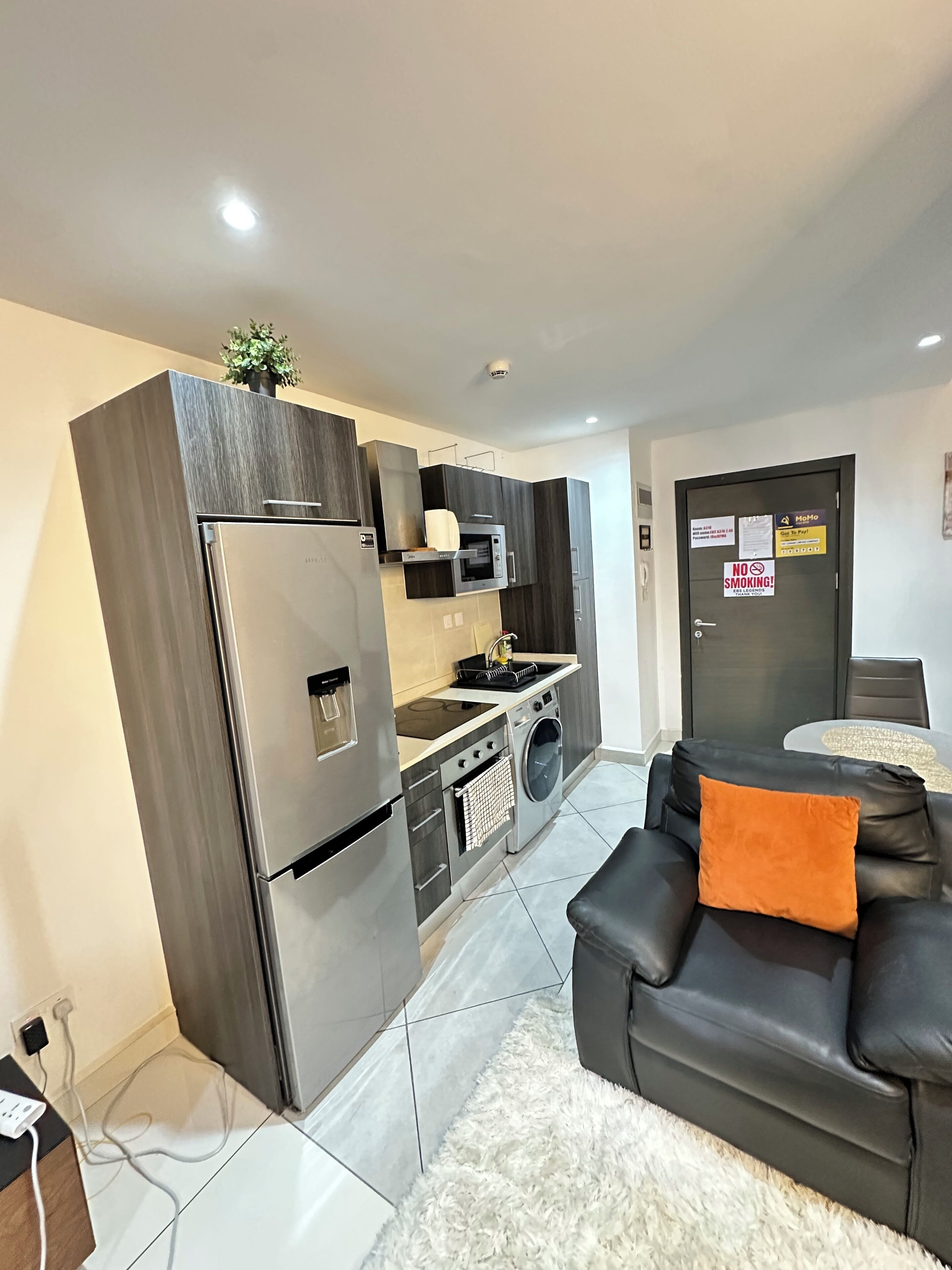 A modern kitchen with a built-in oven, microwave, and washing machine. There is a black leather sofa with an orange pillow and a white fluffy rug.
