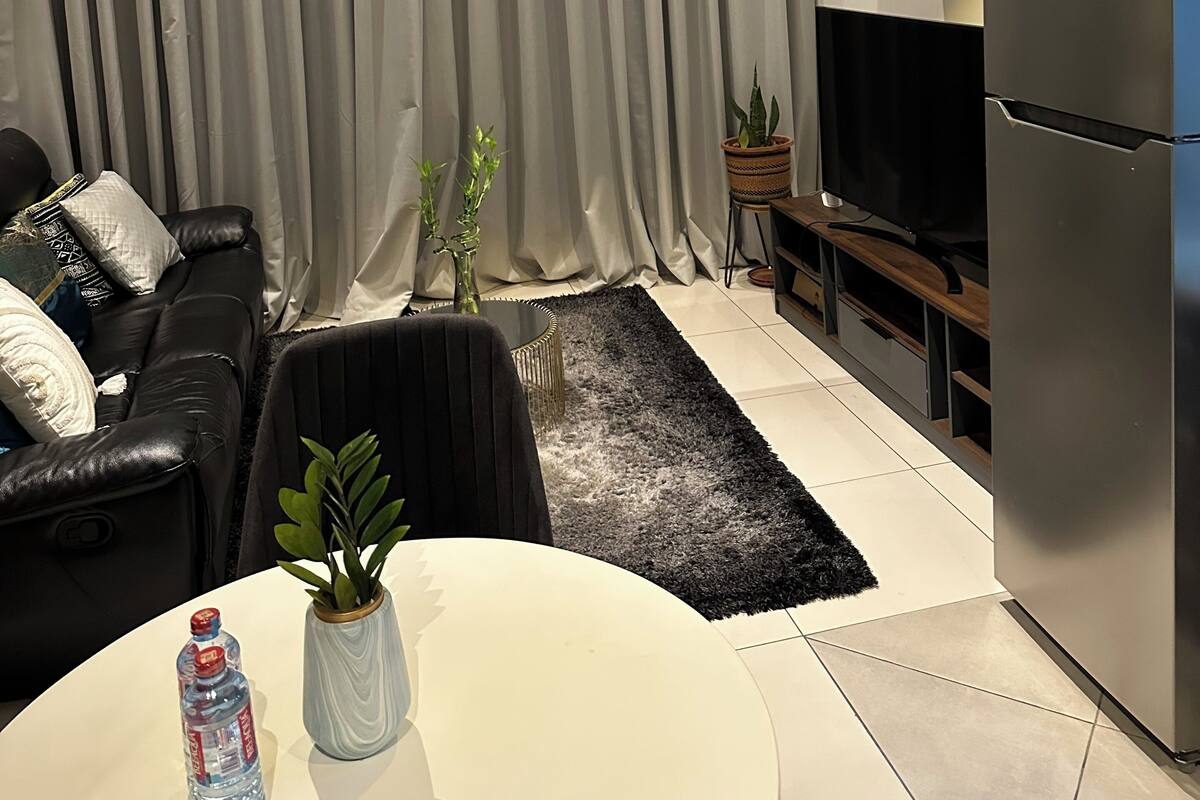 A modern living room with a black leather sofa, a white round table with a plant, a water bottle, and a flat-screen TV.