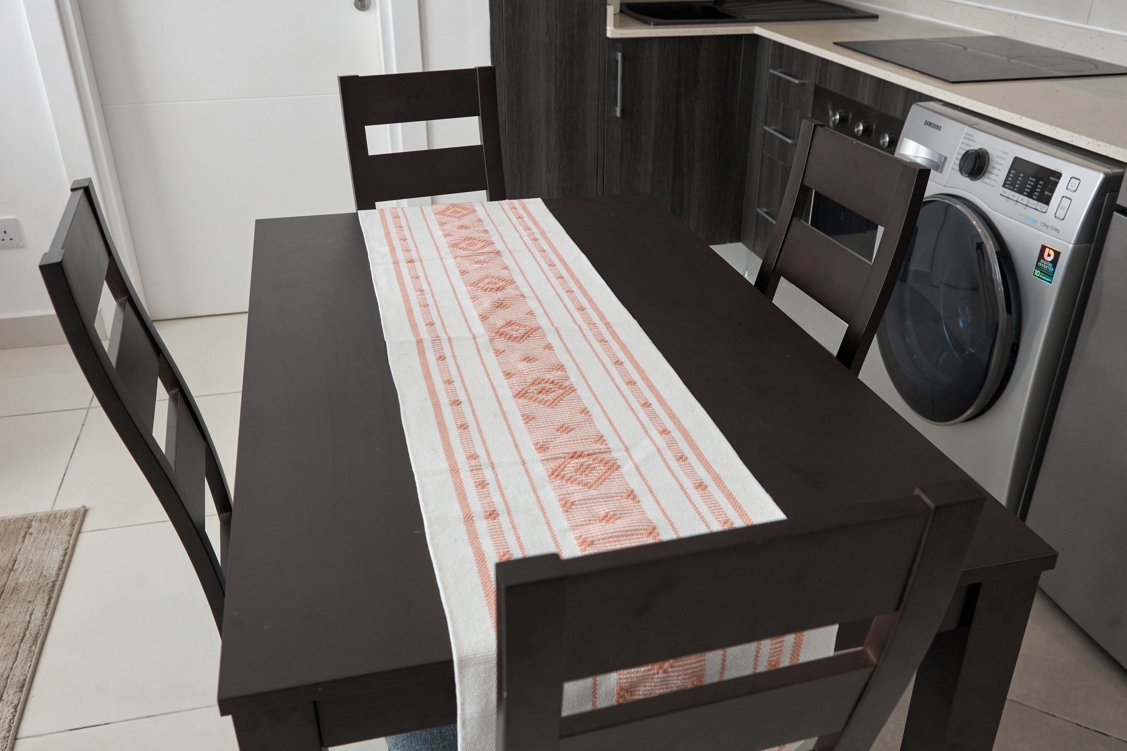 A kitchen with a washing machine, a dining table with a patterned tablecloth, and dark wooden chairs.