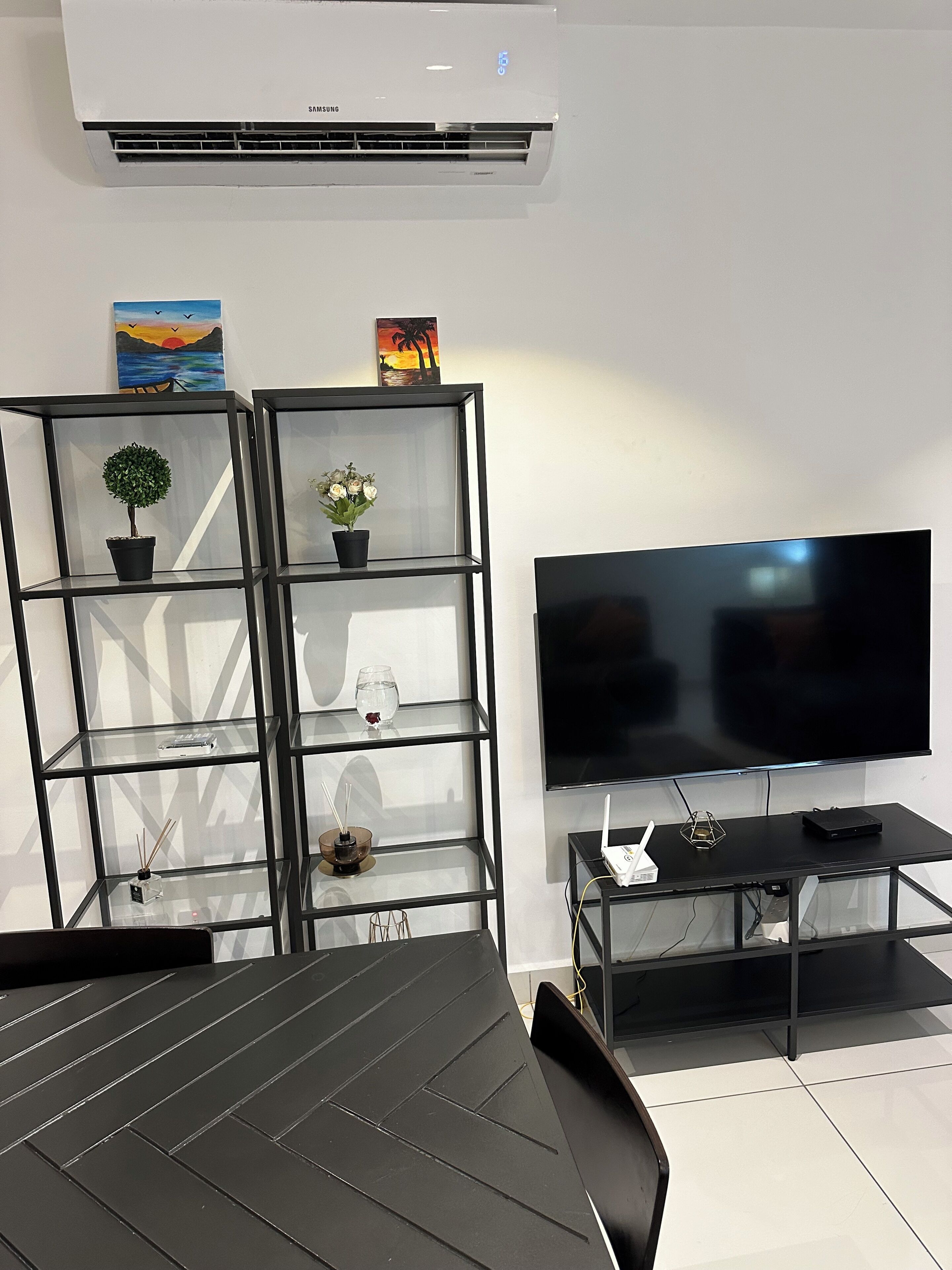 A modern living room with a flat-screen TV, a dining table, and a black sofa. There are two black shelving units with decorative items and a small potted plant.
