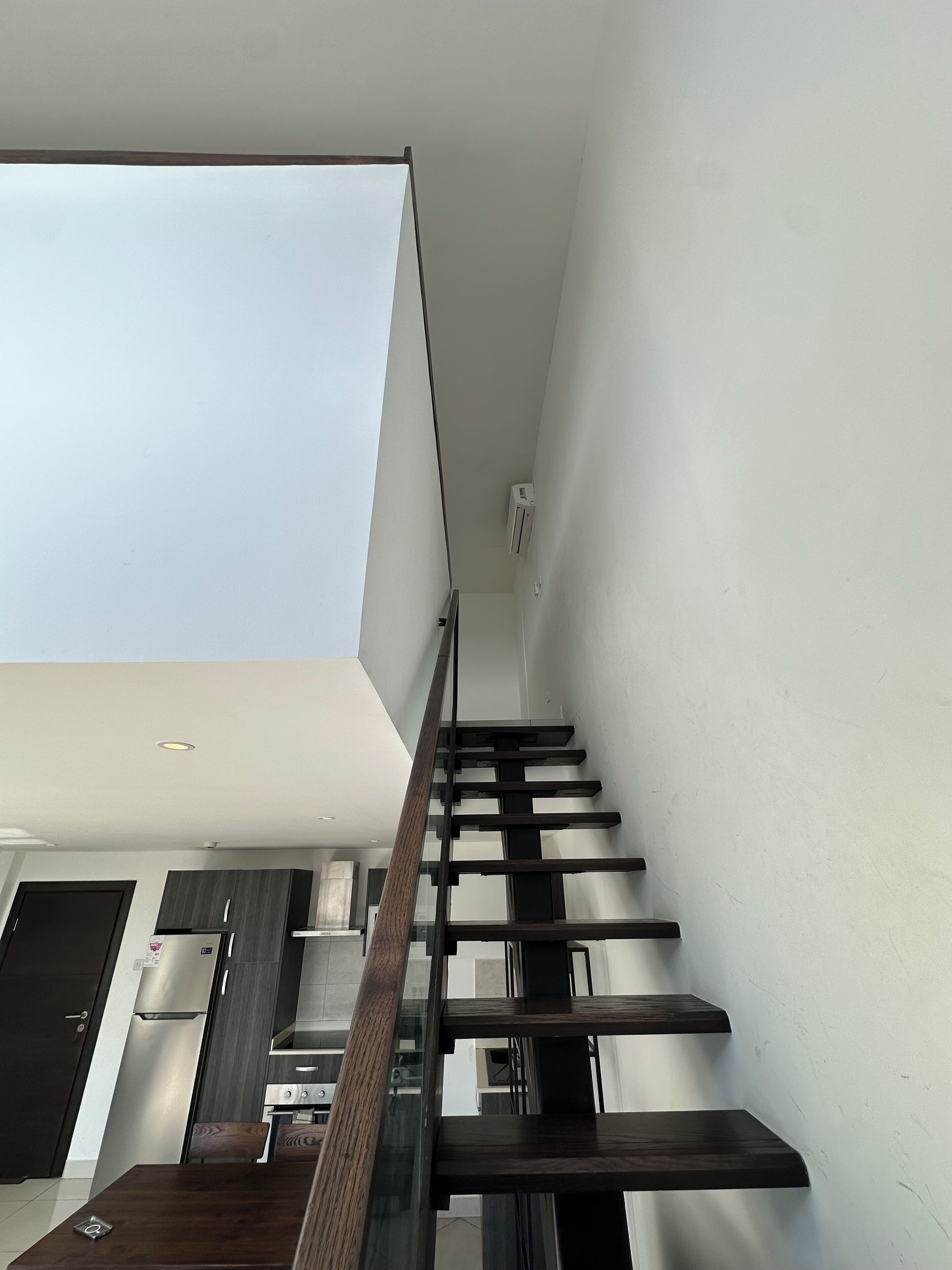 A modern kitchen with dark cabinetry, a stainless steel refrigerator, and a glass staircase leading to a loft area.