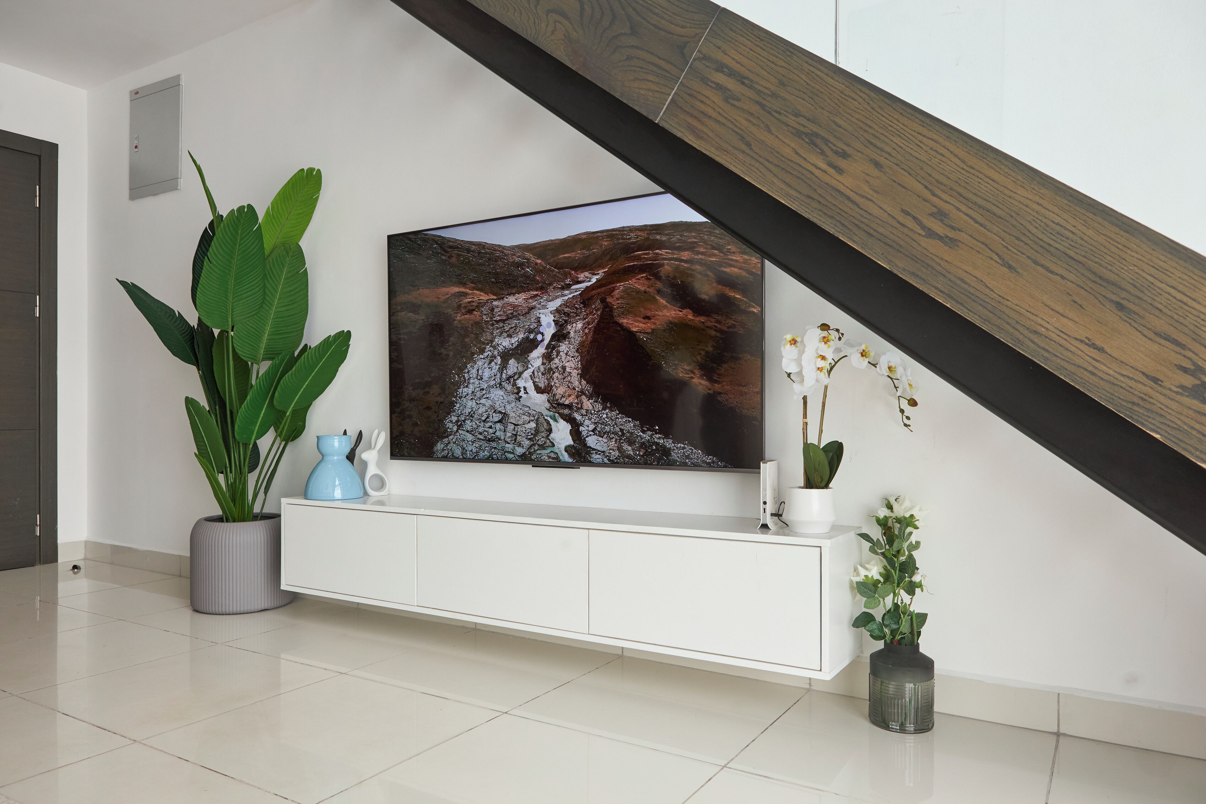 A modern living room with a large flat-screen TV mounted on a white console, a potted plant, and a vase with flowers.